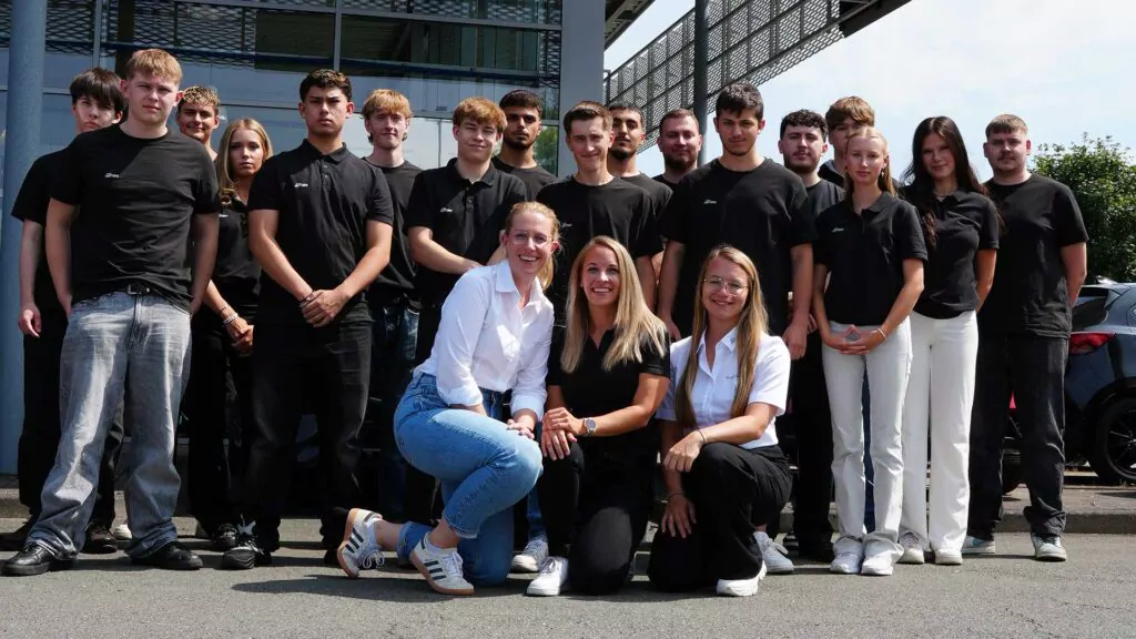 Eine große Gruppe junger Auszubildender in schwarzer Arbeitskleidung posiert lachend im Freien vor einem Gebäude - Gruppenfoto der Azubi-Rallye 2026 der Auto Mattern Gruppe.