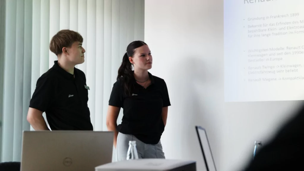 Zwei Auszubildende in schwarzen Hemden stehen neben einer Präsentationsleinwand in einem hellen Schulungsraum - Eindruck von den Onboarding-Tagen der neuen Azubis bei der Auto Mattern Gruppe.