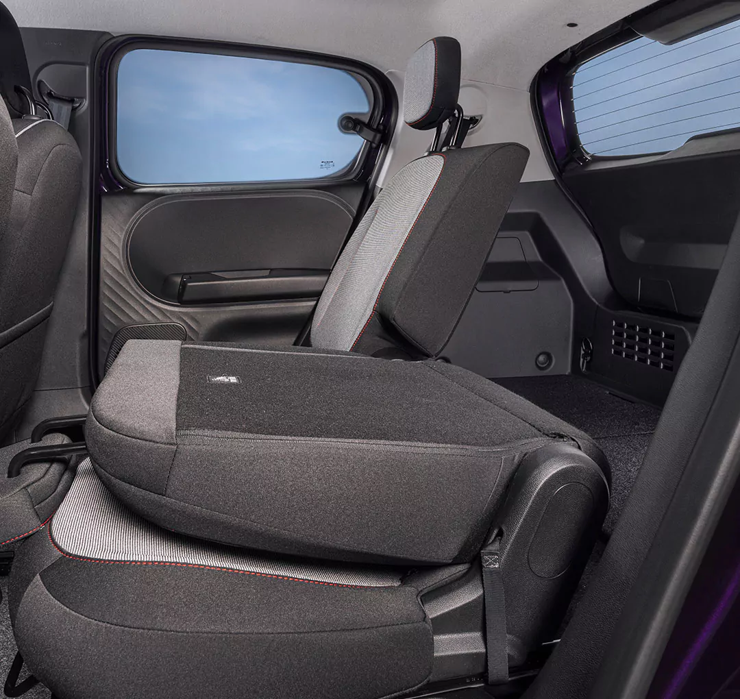 Das Bild zeigt den Rücksitz eines Nissan Pixo, wobei ein Rücksitz umgeklappt ist, um den Laderaum zu vergrößern. Die Polsterung ist dunkel mit hellgrauen Akzenten, und die Autotür sowie das Fenster sind zu sehen.
