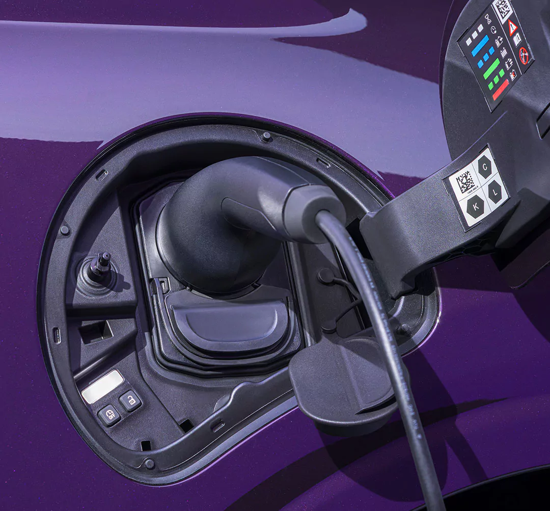 Eine Nahaufnahme der Ladeanschlussbuchse eines Nissan Pixo Elektroautos, an die ein Ladekabel angeschlossen ist. Das Auto ist violett, und auf dem Armaturenbrett sind die Ladeanzeigen zu sehen.