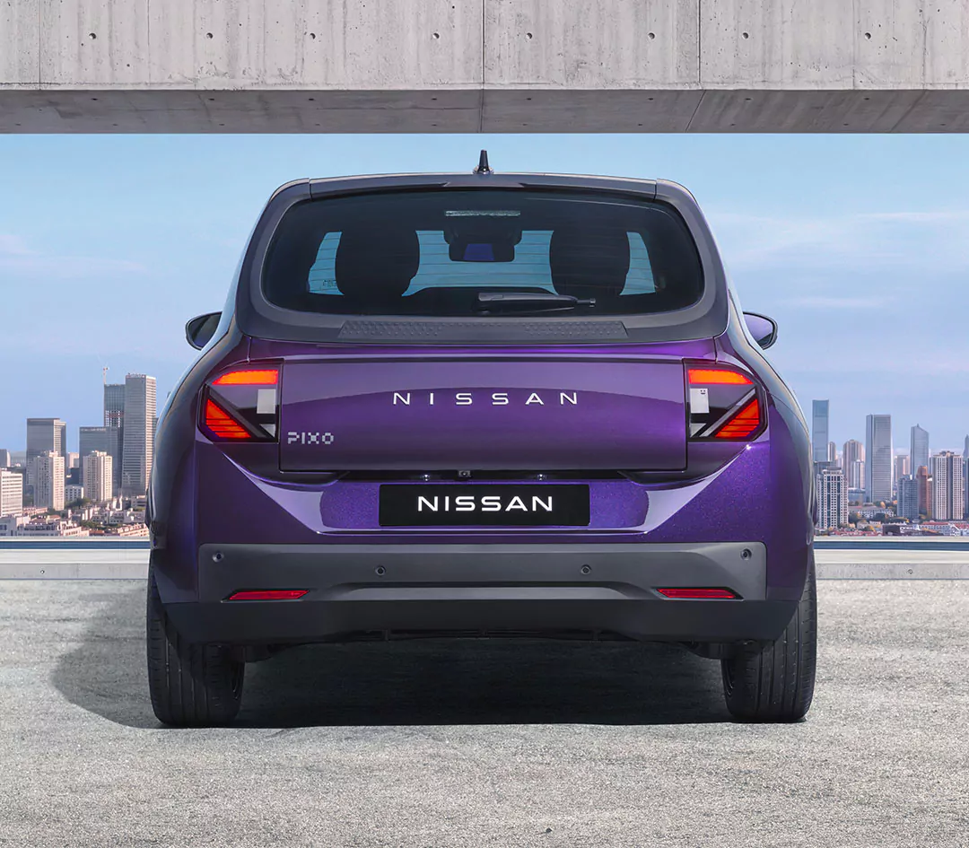 Ein violetter Nissan Pixo ist von hinten zu sehen, geparkt auf einer Betonfläche, im Hintergrund ist die Skyline der Stadt unter einem strahlend blauen Himmel zu erkennen. Das unverwechselbare Design dieses Nissan Pixo hebt sich deutlich von der städtischen Kulisse ab.