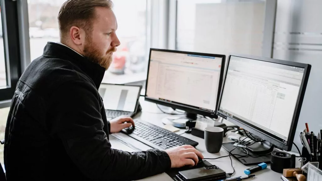 Ein Mann sitzt an einem Schreibtisch und arbeitet an zwei Computermonitoren, auf denen Tabellenkalkulationen und Daten angezeigt werden – typische Aufgaben in einer Ausbildung zum Büromanager. Er wirkt konzentriert und ist umgeben von einer Tastatur, einer Maus, einem Telefon, einer Tasse und Büromaterial.