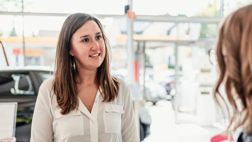 Eine Frau in einer weißen Bluse steht in einem Raum und unterhält sich mit einer anderen Person, die ihr den Rücken zuwendet, über die Ausbildung zum Büromanager. Im hellen Hintergrund sind Autos und große Fenster zu sehen.