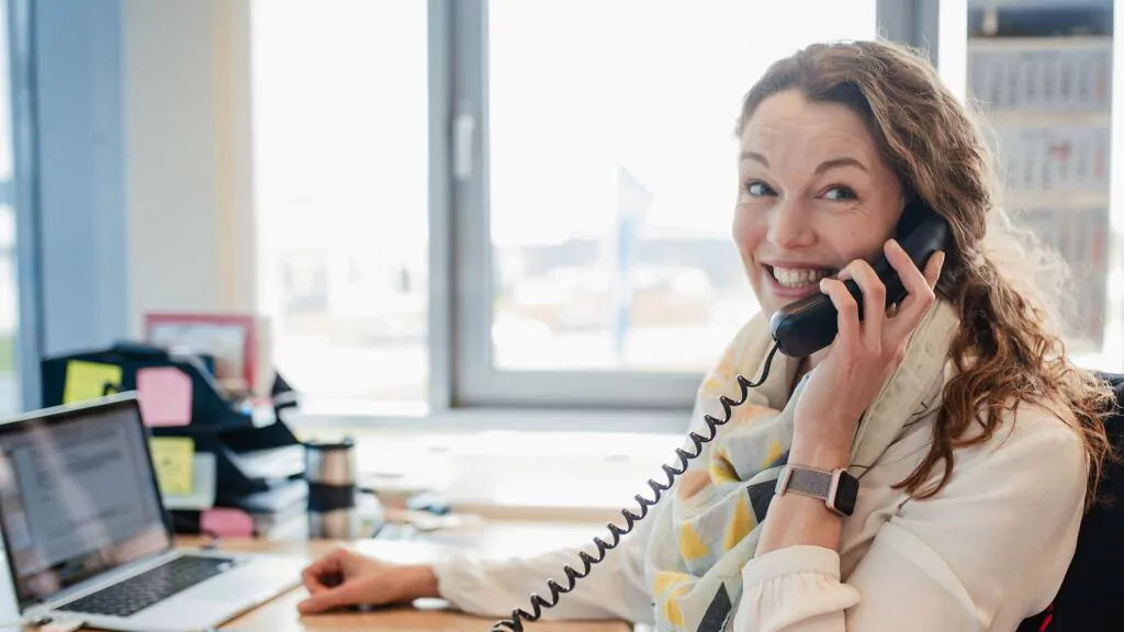 Eine lächelnde Frau telefoniert an ihrem Schreibtisch mit einem Festnetztelefon; daneben sind ein Laptop, Unterlagen und Büromaterial zu sehen – die perfekte Atmosphäre für die Ausbildung zum Büromanager. Sonnenlicht strömt durch ein Fenster hinter ihr herein.