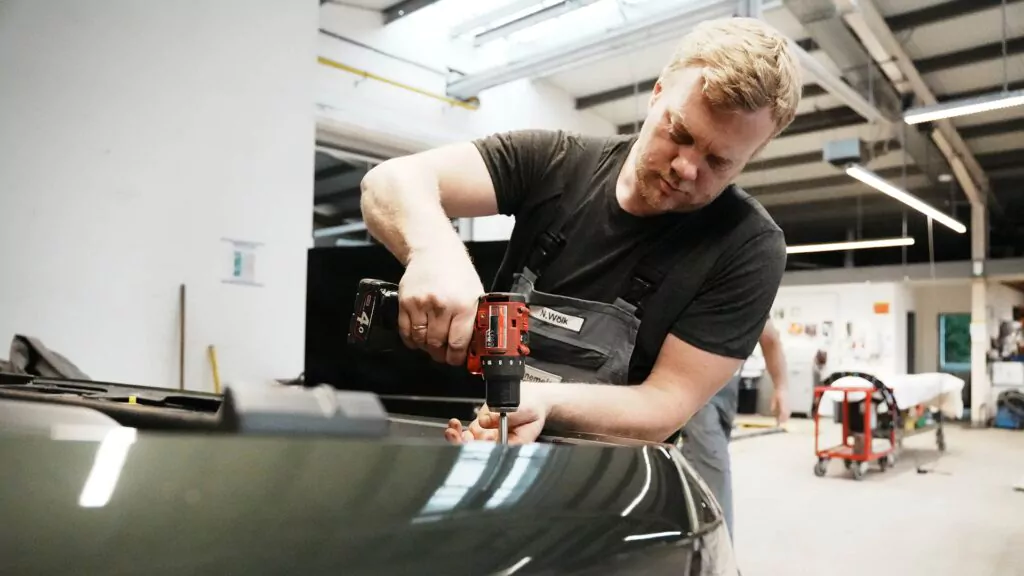 Ein Mann in Arbeitskleidung arbeitet mit einer Bohrmaschine an einem Fahrzeug in einer gut beleuchteten Garage und stellt dabei seine Fähigkeiten aus seiner Ausbildung zum Karosseriebauer unter Beweis. Im Hintergrund sind Werkzeuge und Arbeitsgeräte zu sehen.