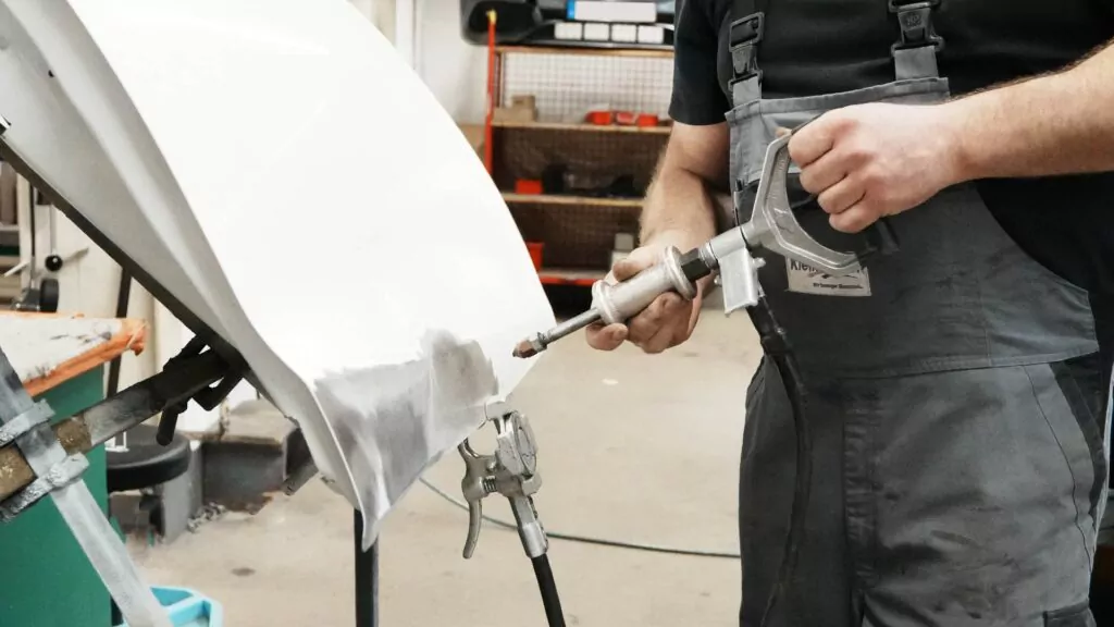 Eine Person in einer Latzhose, die sich möglicherweise in der Ausbildung zum Karosseriebauer befindet, schleift oder poliert mit einem Elektrowerkzeug ein weißes Autoteil, das in einer Werkstatt auf einem Ständer befestigt ist.
