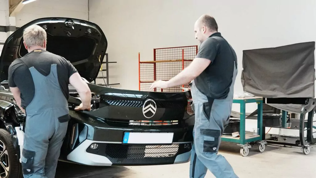 Zwei Mechaniker in grauen Overalls, die möglicherweise eine Ausbildung zum Karosseriebauer absolvieren, arbeiten in einer gut beleuchteten Kfz-Werkstatt an einem schwarzen Citroën mit geöffneter Motorhaube.