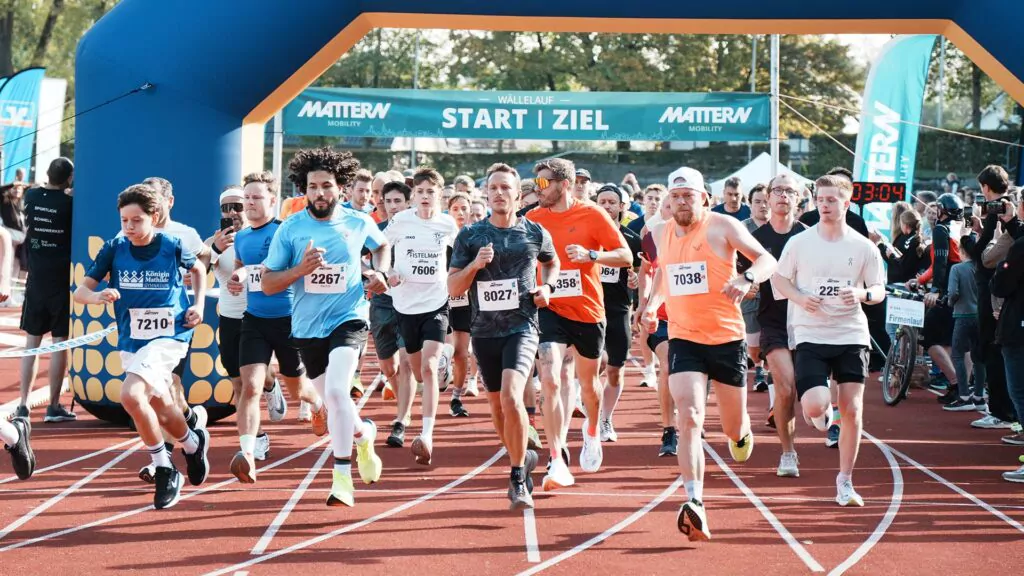 Eine Gruppe von Läufern in Sportbekleidung, die an einem Rennen auf einer Außenbahn teilnehmen und unter einem Start- und Zielbogen hindurchlaufen.